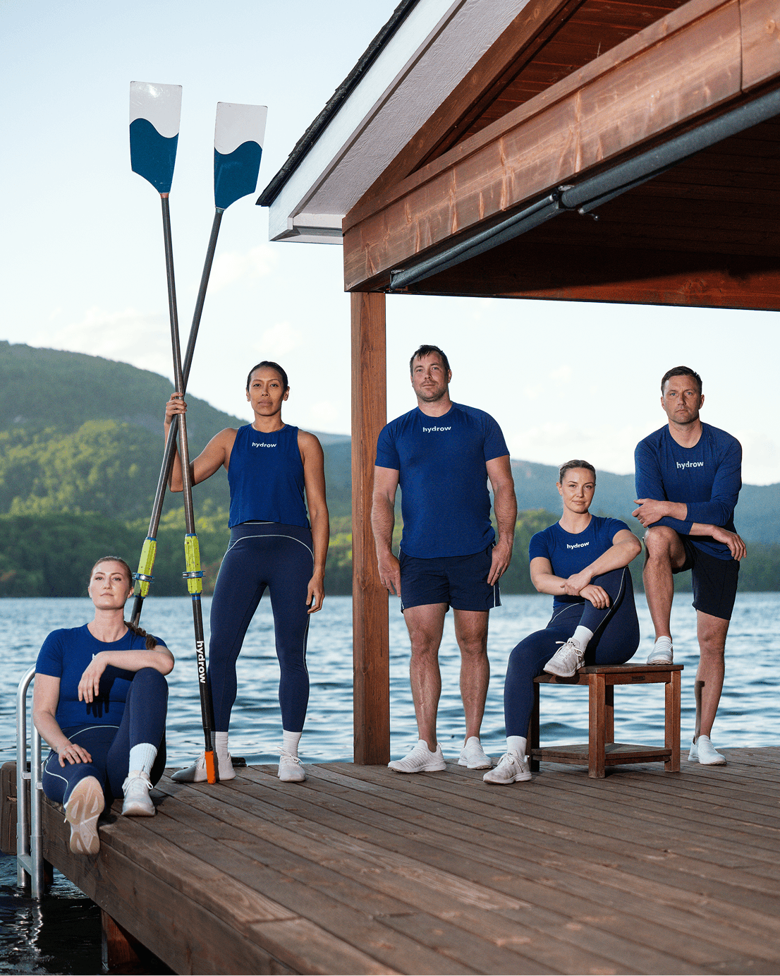 Hydrow athletes standing together on a lakeside dock holding oars